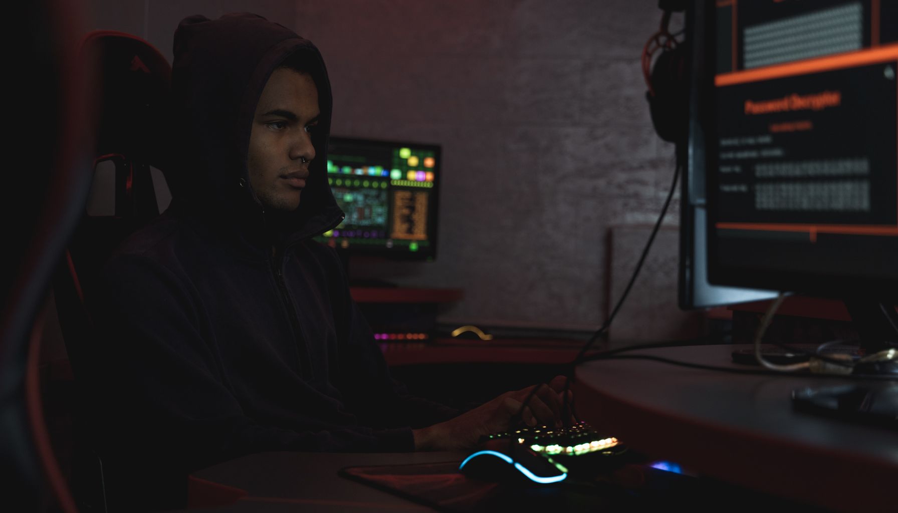 A hacker in a hoodie working in a dimly lit room, focusing on cyber security tasks on multiple monitors.