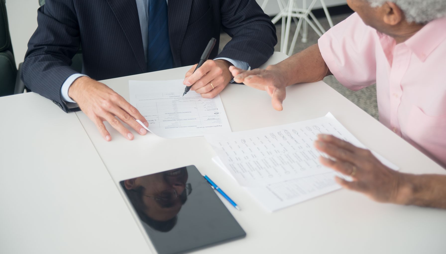 A business meeting between an elderly client and a consultant discussing documents at an office table.