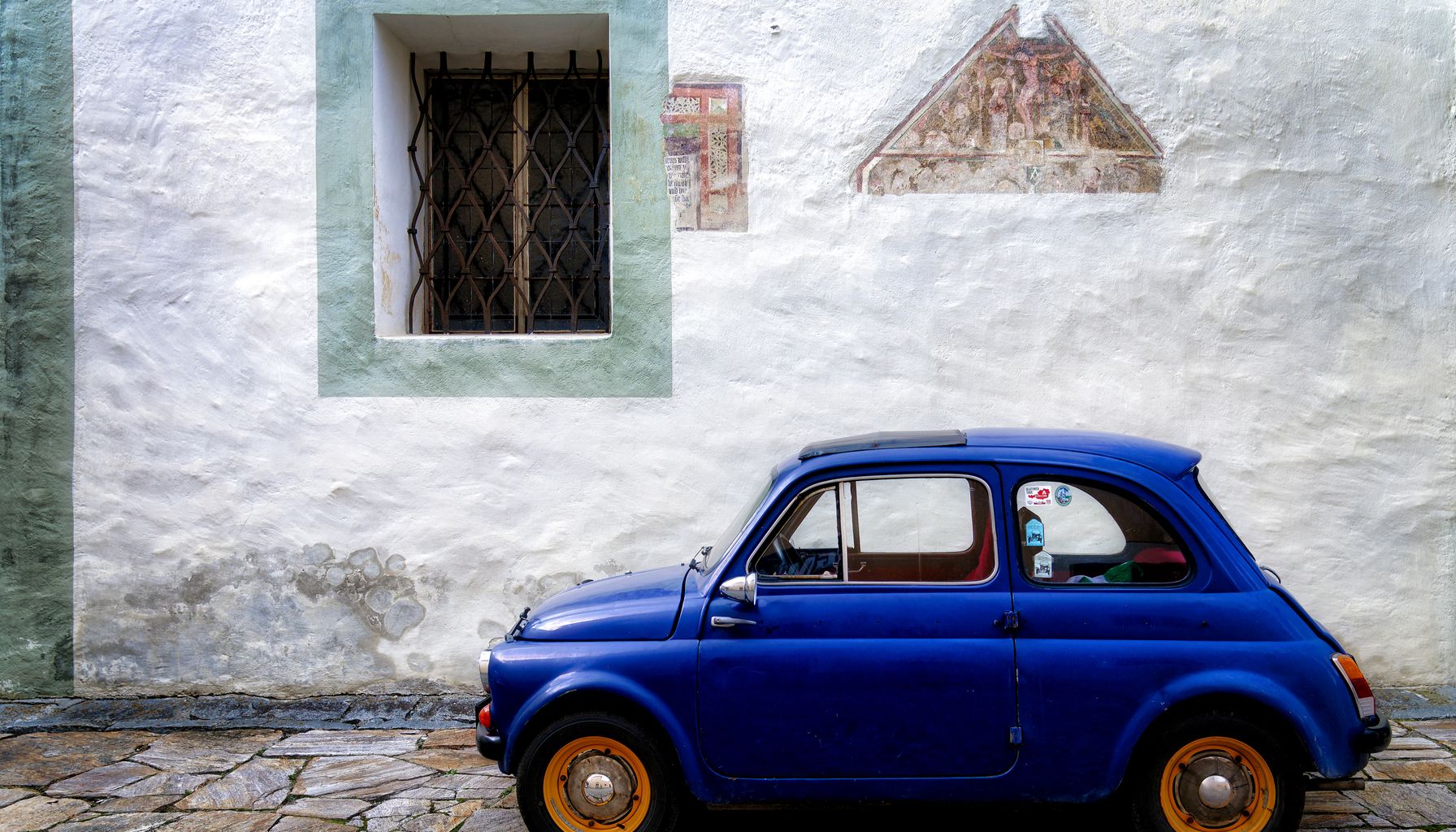 A classic blue car parked by a textured wall with historical architecture.
