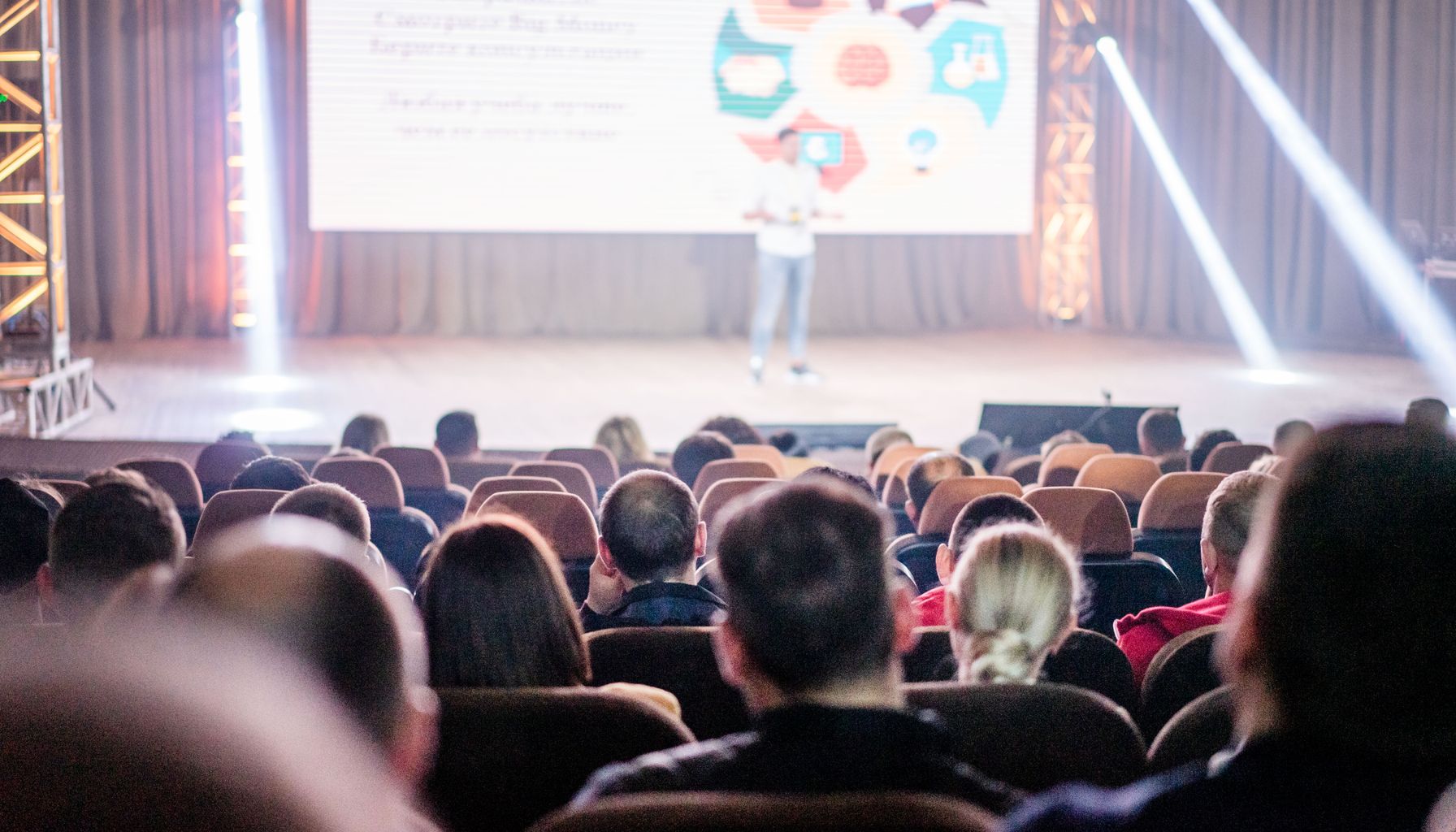 Audience attentively watching a presentation in a conference setting, with stage lights and speaker visible.