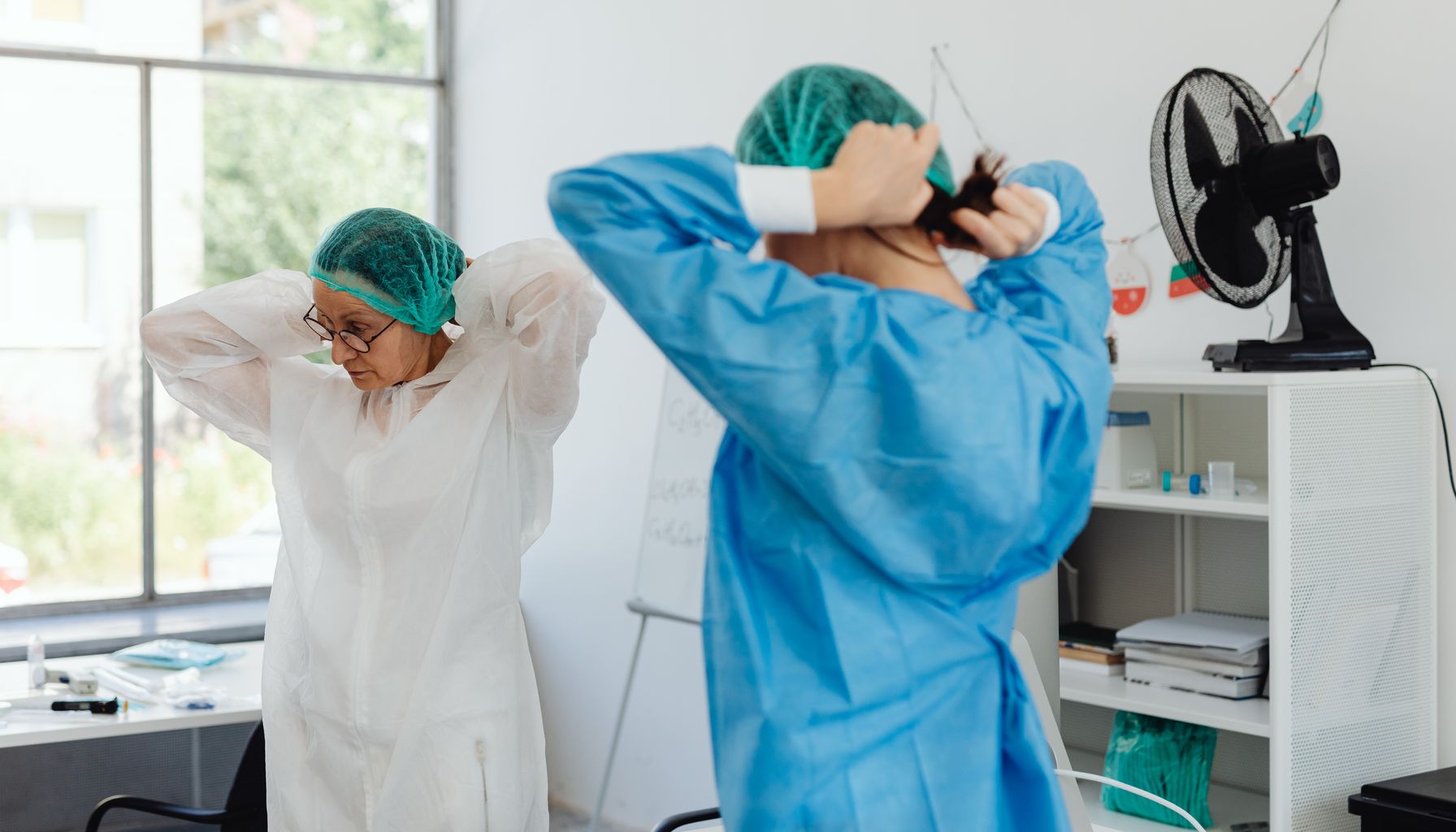 Two healthcare workers in scrubs and hair nets getting ready in a hospital room.