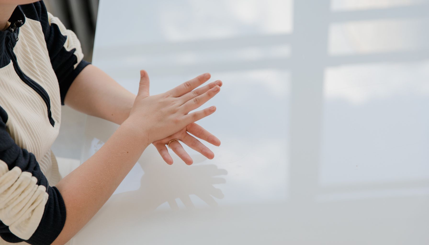 An adult gesturing hands on a reflective white table indoors, with soft sunlight.