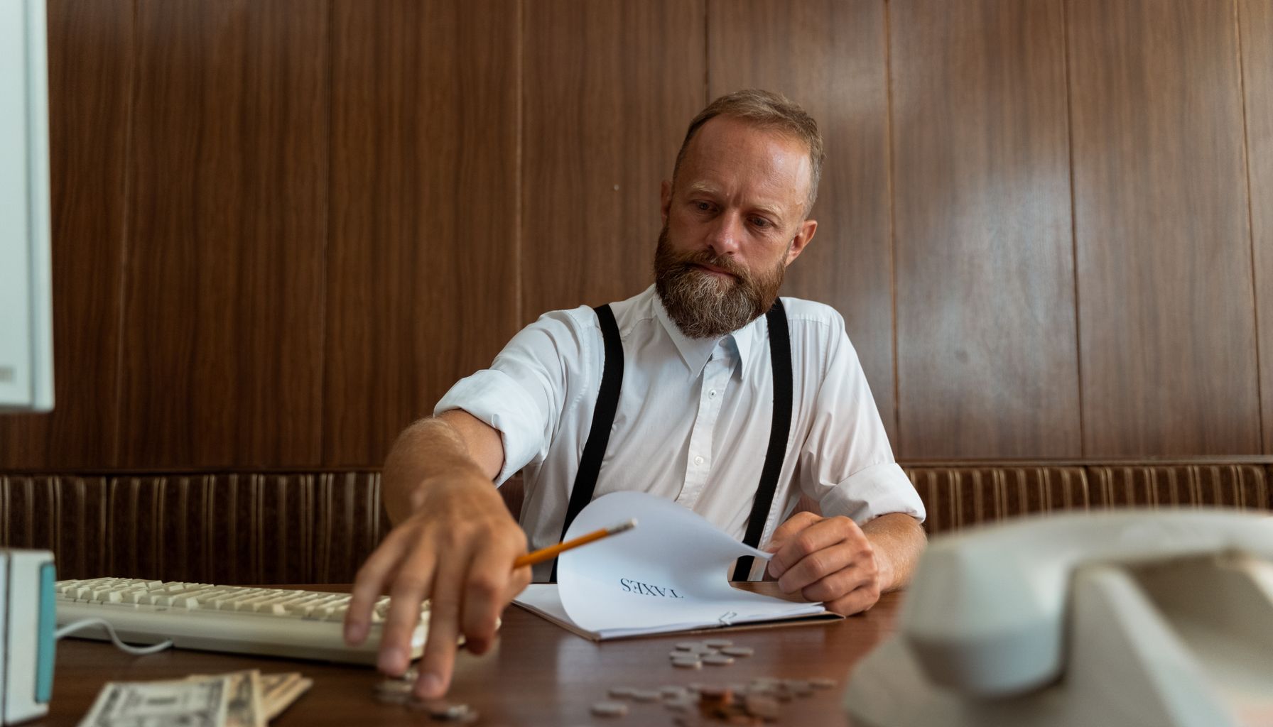 Bearded man in vintage office calculating finances with retro telephone and typewriter.
