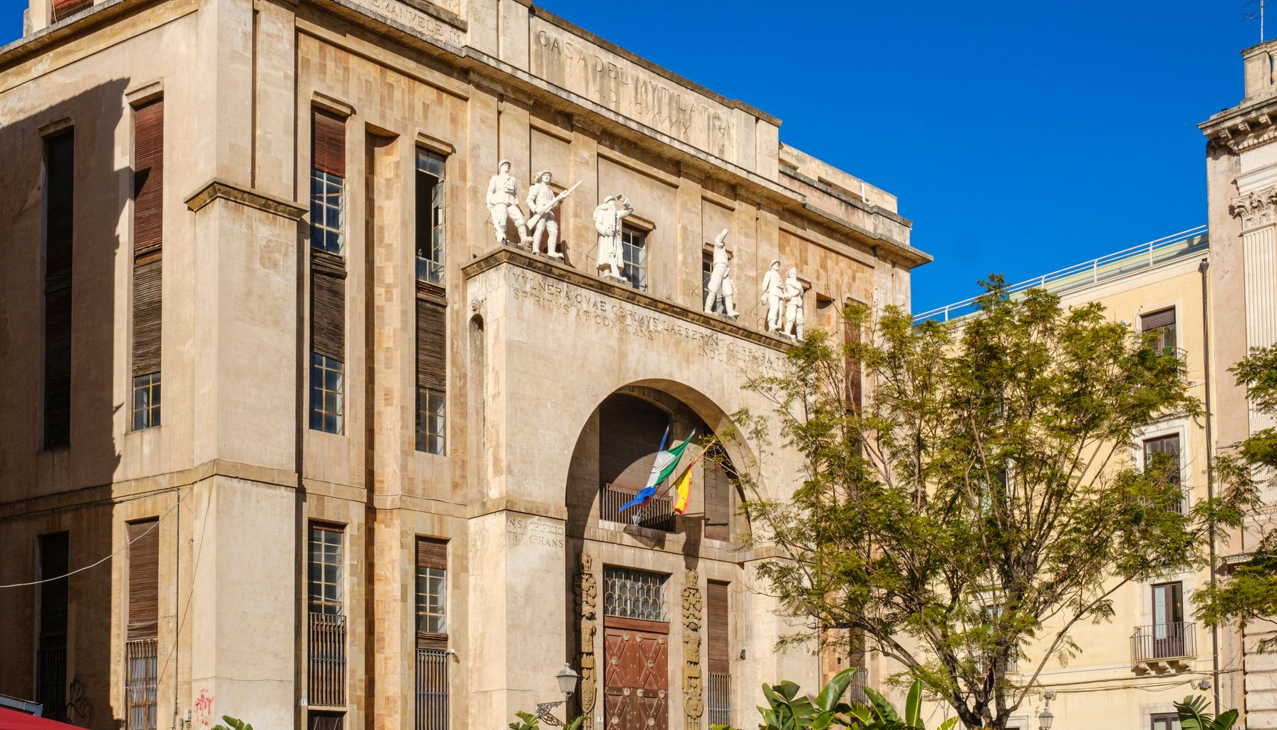 Photograph of a historic building in Catania, Sicily with statues and flags on a sunny day.