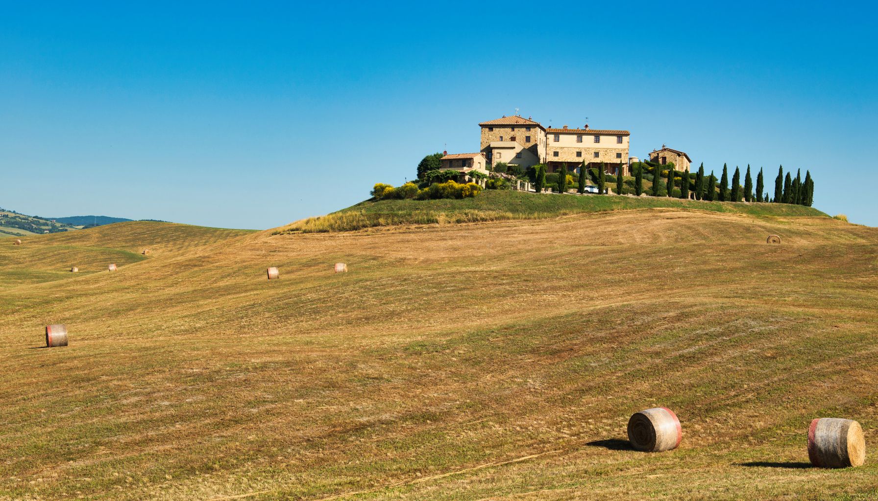 A picturesque Tuscan farmhouse atop rolling hills surrounded by hay bales under a clear summer sky.