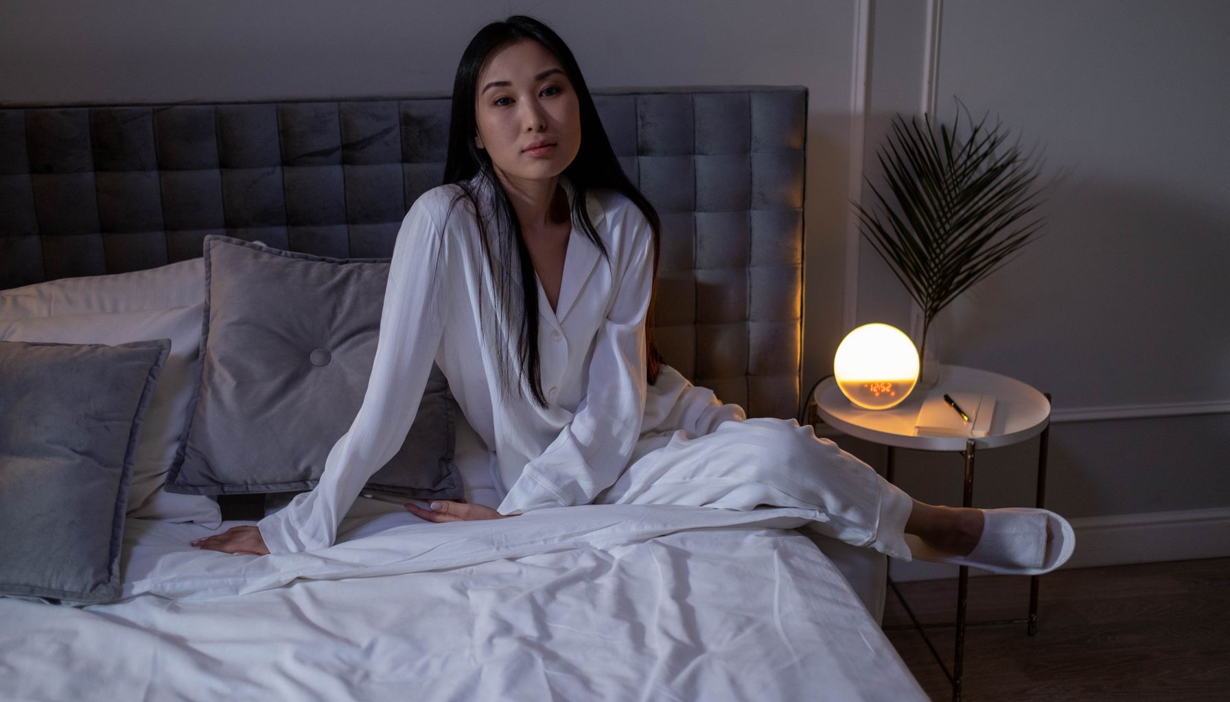 A woman in white sleepwear sitting on a modern bed in a cozy bedroom.