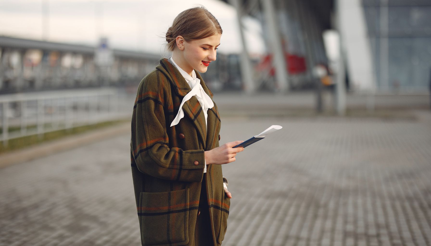 Smiling female passenger wearing trendy plaid coat and white blouse checking passport and ticket standing on pavement near contemporary building of airport